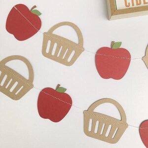 Peut inclure: Une guirlande en papier rouge et marron avec des pommes et des paniers. La guirlande est enfil&eacute;e sur un fil blanc.
