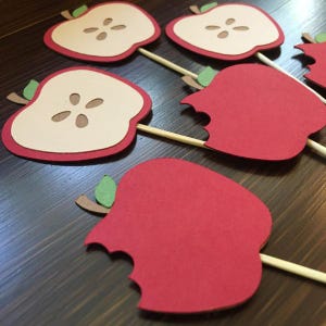 May include: A set of six red apple-shaped cupcake toppers. The toppers are made of paper and have a green leaf on top. Some of the apples have a bite taken out of them, while others are whole. The apples are on wooden skewers.