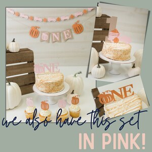 May include: A first birthday cake with a pumpkin theme. The cake is decorated with a brown and orange 'ONE' topper and a pumpkin. There are also cupcakes with pumpkin toppers. The cake is on a white cake stand. The background is a wooden crate with pumpkins and a banner that says 'ONE' in brown and pink.
