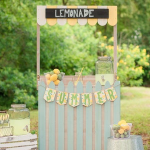 May include: A wooden lemonade stand painted in light blue and white with a sign that says "Lemonade" and a banner that says "Summer". There are two glass jars, one with lemonade and the other empty, and two baskets of lemons.