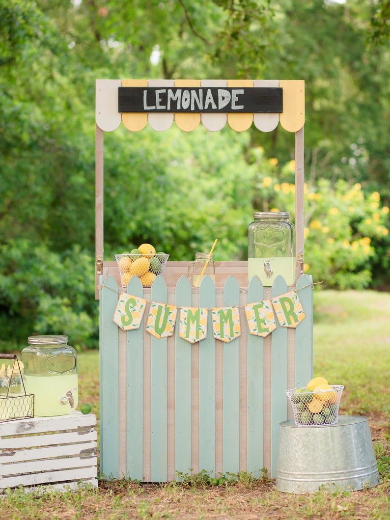 May include: A wooden lemonade stand painted in light blue and white with a sign that says "Lemonade" and a banner that says "Summer". There are two glass jars, one with lemonade and the other empty, and two baskets of lemons.