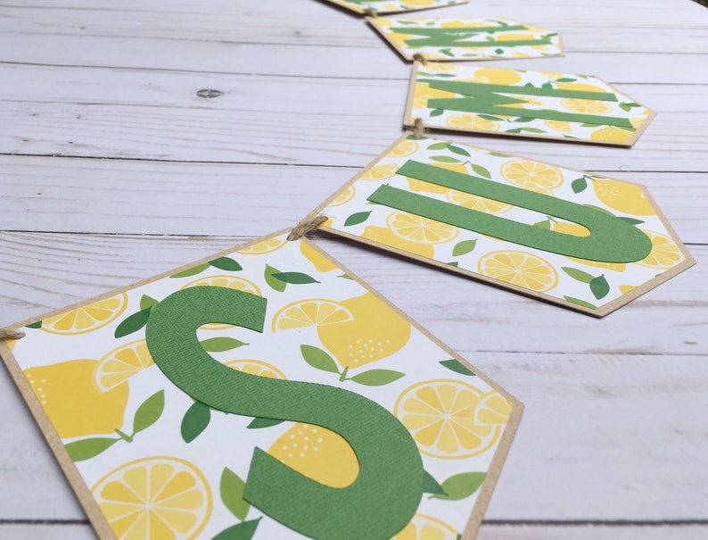 May include: A summer banner with the word "SUMMER" spelled out in green letters on a white background with a lemon pattern. The banner is made of brown cardstock and is strung together with twine.
