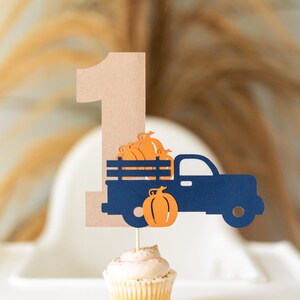 May include: A brown cardboard number one with a blue truck carrying orange pumpkins. The truck is on a cupcake with brown frosting.