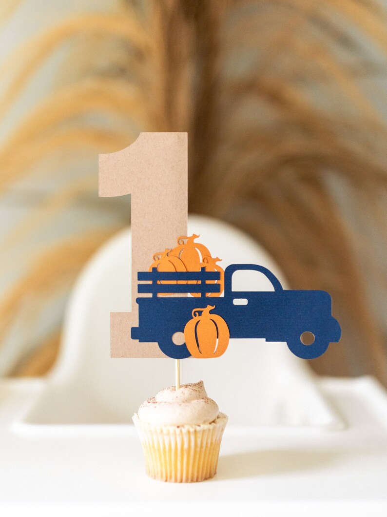 May include: A brown cardboard number one with a blue truck carrying orange pumpkins. The truck is on a cupcake with brown frosting.