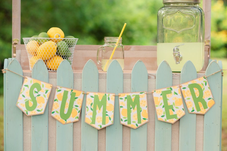 May include: A lemonade stand with a green picket fence and a banner that says "SUMMER" in green letters. The banner has a lemon and leaf pattern. There is a pitcher of lemonade, a glass of lemonade, and a basket of lemons.