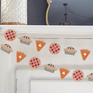 May include: A festive garland with alternating pie shapes in red, orange, and brown. The garland is made of paper and is strung together with white string.