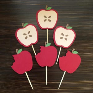 May include: Six red apple cupcake toppers, some with a bite taken out, and some with a brown center showing the inside of the apple. The toppers are on wooden sticks.