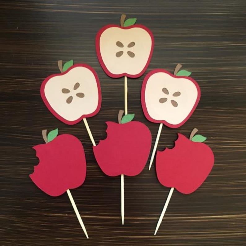 May include: Six red apple cupcake toppers, some with a bite taken out, and some with a brown center showing the inside of the apple. The toppers are on wooden sticks.