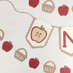 Peut inclure: Une banni&egrave;re &agrave; th&egrave;me pomme rouge et blanche avec la lettre "N" et des paniers. La banni&egrave;re est enfil&eacute;e avec de la ficelle et comporte des pommes rouges et des paniers.