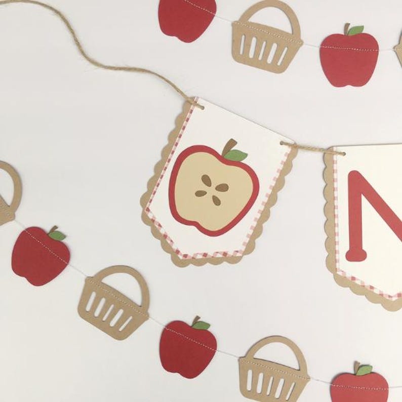 Peut inclure: Une banni&egrave;re &agrave; th&egrave;me pomme rouge et blanche avec la lettre "N" et des paniers. La banni&egrave;re est enfil&eacute;e avec de la ficelle et comporte des pommes rouges et des paniers.