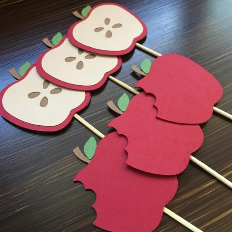 May include: Set of six red and cream paper apple cupcake toppers. Three toppers feature a whole apple with a bite taken out of it. The other three toppers feature a slice of apple with a brown seed in the center.