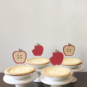 May include: Four mini apple pies on white cake stands with red apple toppers. The apple toppers have a bite taken out of them.