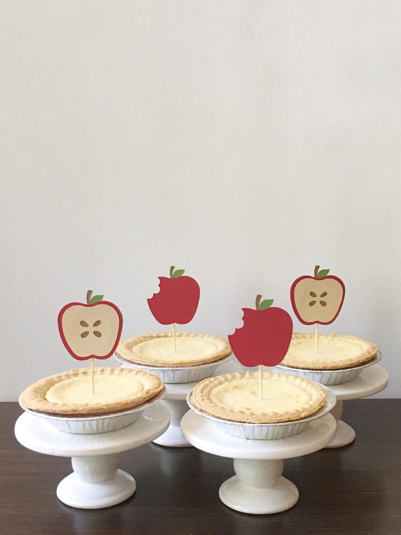May include: Four mini apple pies on white cake stands with red apple toppers. The apple toppers have a bite taken out of them.
