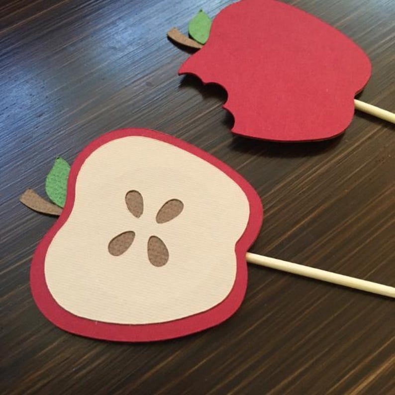 May include: Two red paper apple cutouts on wooden sticks. One apple is whole and the other has a bite taken out of it. The whole apple has a brown paper center with five seeds.