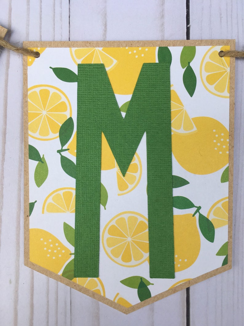May include: A green letter "M" on a white background with a lemon pattern. The letter is cut out of green cardstock and is attached to a brown cardstock banner.