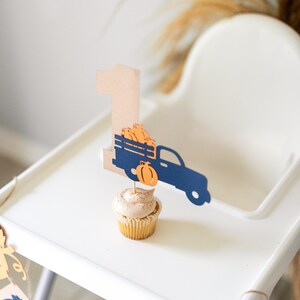 May include: A white high chair with a brown and blue paper truck and pumpkin topper on a cupcake. The truck is carrying pumpkins and the number one is on the side of the truck. There is a banner with pumpkins hanging from the chair.