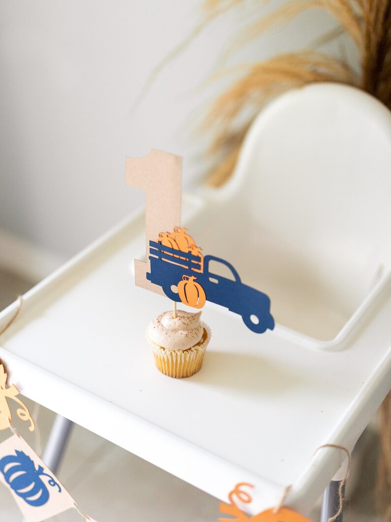 May include: A white high chair with a brown and blue paper truck and pumpkin topper on a cupcake. The truck is carrying pumpkins and the number one is on the side of the truck. There is a banner with pumpkins hanging from the chair.