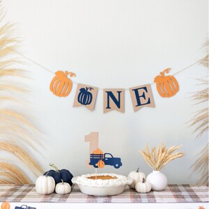 May include: A brown burlap banner with the word "ONE" in navy blue letters and orange pumpkin cutouts. A small, brown paper number "1" with a blue truck carrying orange pumpkins is on a white cake with a white pie crust. There are orange pumpkins and blue trucks on a white table with a plaid tablecloth.