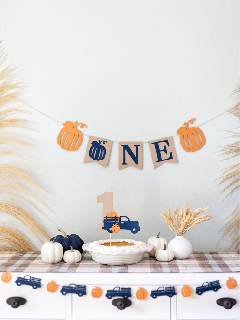 May include: A brown burlap banner with the word "ONE" in navy blue letters and orange pumpkin cutouts. A small, brown paper number "1" with a blue truck carrying orange pumpkins is on a white cake with a white pie crust. There are orange pumpkins and blue trucks on a white table with a plaid tablecloth.