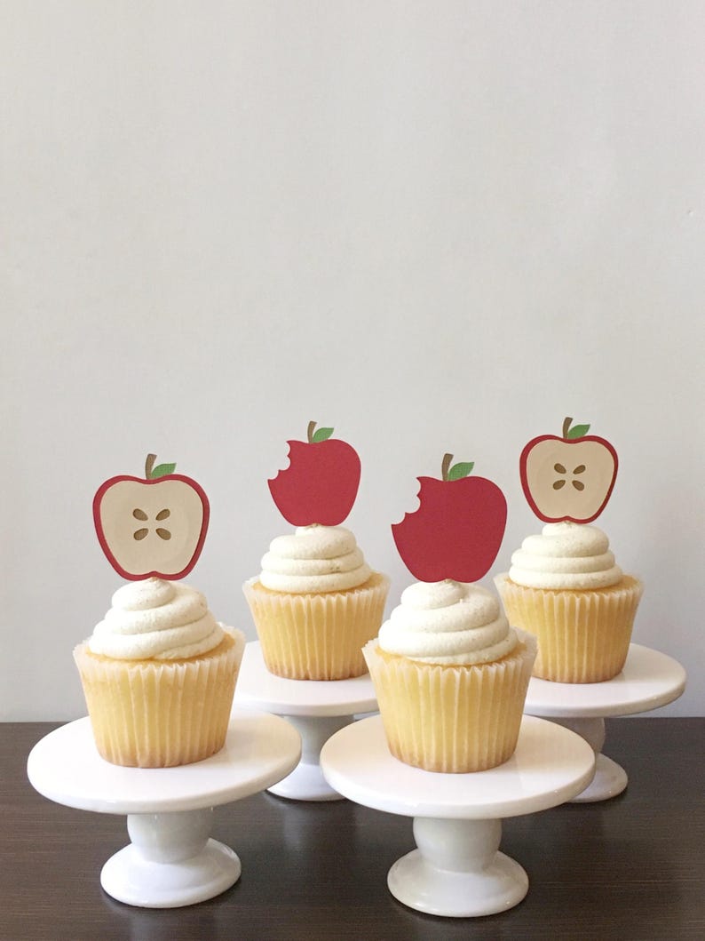 May include: Four white cupcake stands with yellow cupcakes topped with white frosting and red apple cupcake toppers. The apple toppers have a bite taken out of them.