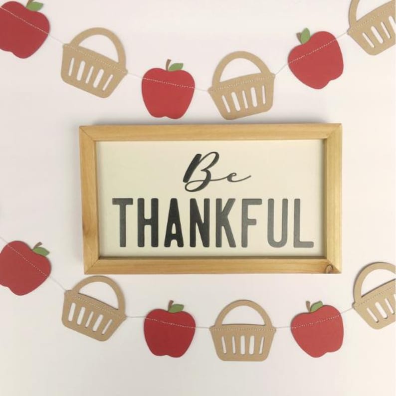 Peut inclure: Une guirlande en papier avec des d&eacute;coupes de pommes rouges et de paniers bruns. La guirlande est enfil&eacute;e sur un fil blanc. Un panneau en bois avec le texte "Be Thankful" est en arri&egrave;re-plan.