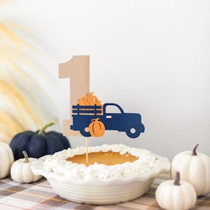 May include: A brown paper number one with a blue truck carrying orange pumpkins on top of a pumpkin pie with whipped cream. The pie is on a plaid tablecloth with white pumpkins around it.