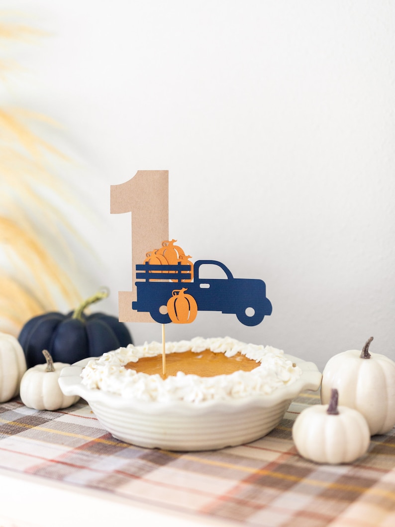 May include: A brown paper number one with a blue truck carrying orange pumpkins on top of a pumpkin pie with whipped cream. The pie is on a plaid tablecloth with white pumpkins around it.