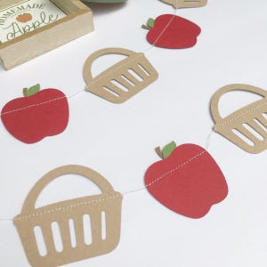 Peut inclure: Une guirlande en papier rouge et marron avec des pommes et des paniers altern&eacute;s. La guirlande est enfil&eacute;e sur un fil blanc. Les pommes sont rouges avec des tiges et des feuilles vertes. Les paniers sont marrons avec une bordure blanche cousue et une poign&eacute;e blanche cousue.