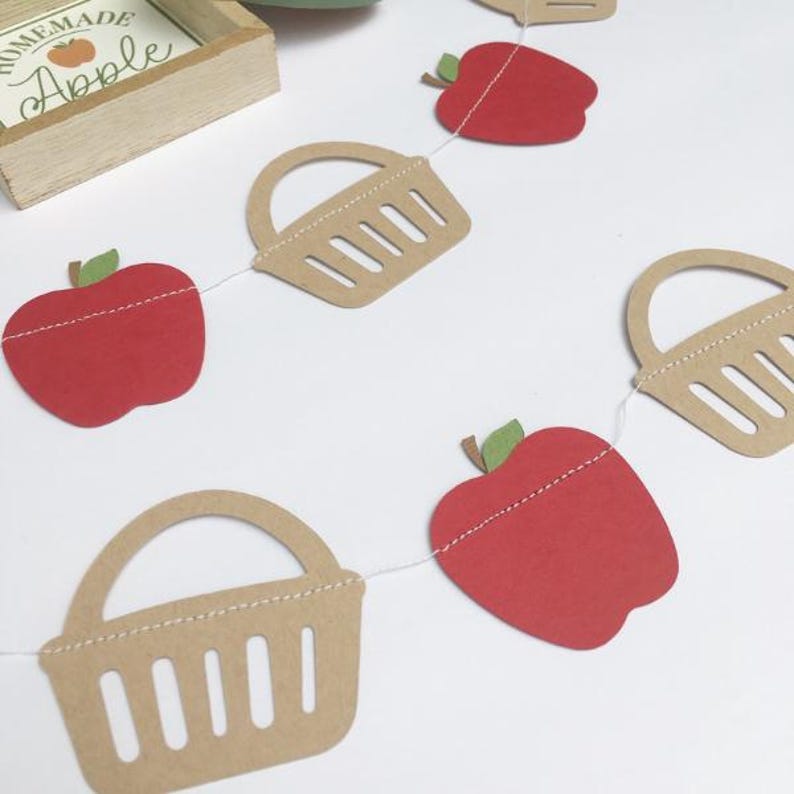 Peut inclure: Une guirlande en papier rouge et marron avec des pommes et des paniers altern&eacute;s. La guirlande est enfil&eacute;e sur un fil blanc. Les pommes sont rouges avec des tiges et des feuilles vertes. Les paniers sont marrons avec une bordure blanche cousue et une poign&eacute;e blanche cousue.