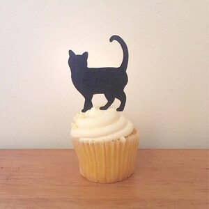 May include: A cupcake with a light-colored paper liner and a swirl of white frosting topped with a black, flat, silhouette-style cutout of a standing cat with its tail curved upward. The cat cutout is positioned vertically on the frosting.