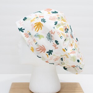 Può includere: Cappello chirurgico bianco con un motivo botanico colorato. Il cappello presenta vari disegni di foglie e fiori nei toni del verde, giallo, arancione e rosa. Le parole "Powerful" e "Strong" sono stampate.