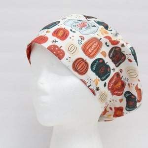 May include: A white surgical scrub cap featuring a repeating pattern of colorful pumpkins, leaves, and floral designs. The pumpkins are in shades of orange, red, and green, with accents of blue and yellow. The cap is designed to fit comfortably.