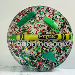 May include: A round, clear resin coaster with a colorful, glittery design. It features a green Lego block, a green eraser, a yellow Crayola crayon, and white letter beads spelling "Miss Becker".