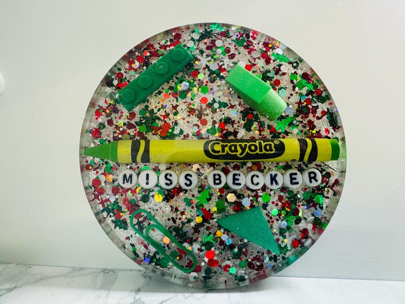 May include: A round, clear resin coaster with a colorful, glittery design. It features a green Lego block, a green eraser, a yellow Crayola crayon, and white letter beads spelling "Miss Becker".