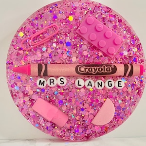 May include: Pink glitter resin coaster with a pink crayon, pink eraser, pink paper clips, and a pink Lego block. The coaster also has the words "Mrs. Lange" in white letters.