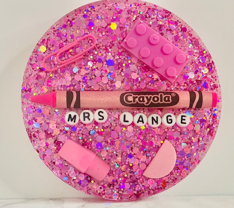 May include: Pink glitter resin coaster with a pink crayon, pink eraser, pink paper clips, and a pink Lego block. The coaster also has the words "Mrs. Lange" in white letters.