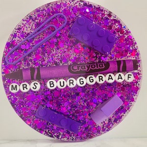 May include: Purple glitter resin coaster with a purple crayon, purple plastic letters spelling "Mrs Burgraaff", and purple plastic building blocks.