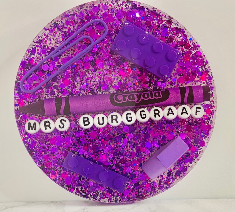 May include: Purple glitter resin coaster with a purple crayon, purple plastic letters spelling "Mrs Burgraaff", and purple plastic building blocks.