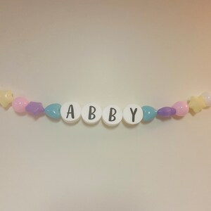 May include: A white beaded garland with the name "ABBY" spelled out in black letters. The garland is decorated with pastel pink, purple, and blue beads.