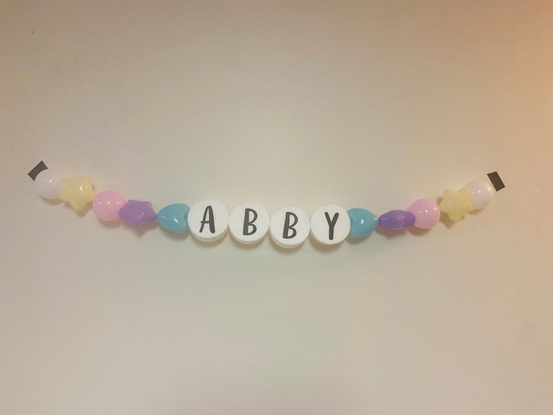 May include: A white beaded garland with the name "ABBY" spelled out in black letters. The garland is decorated with pastel pink, purple, and blue beads.