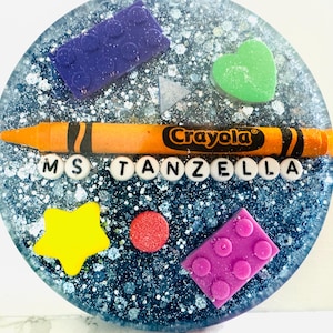 May include: A blue and silver glitter resin coaster with a yellow star, red circle, green heart, purple and pink Lego blocks, an orange crayon with the word "Crayola" on it, and the letters "MS TANZELLA" in white.