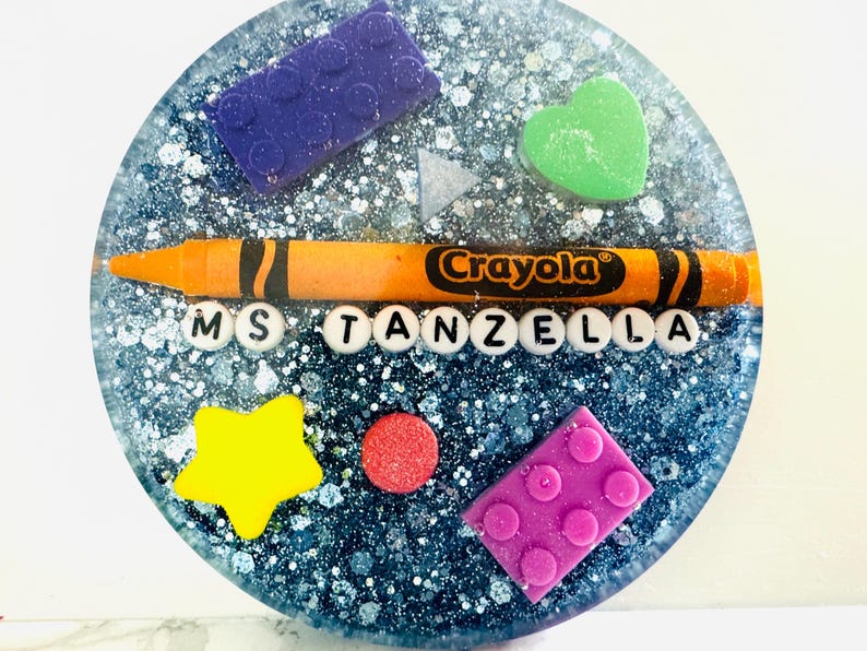 May include: A blue and silver glitter resin coaster with a yellow star, red circle, green heart, purple and pink Lego blocks, an orange crayon with the word "Crayola" on it, and the letters "MS TANZELLA" in white.