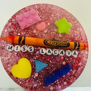 May include: A pink and red glitter resin coaster with a Crayola crayon, a heart, a star, a triangle, and two Lego blocks. The coaster also has the letters "MISS LACAVA" in white.