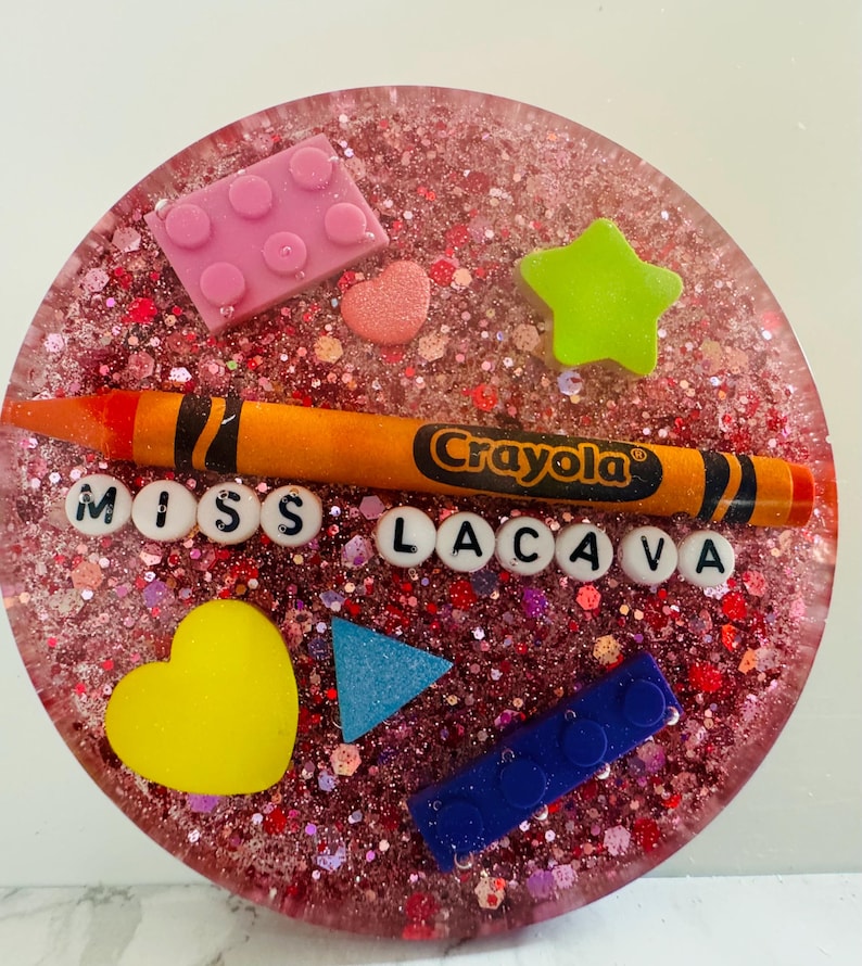 May include: A pink and red glitter resin coaster with a Crayola crayon, a heart, a star, a triangle, and two Lego blocks. The coaster also has the letters "MISS LACAVA" in white.