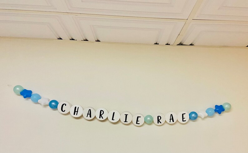 May include: A white banner with the name "CHARLIE RAE" spelled out in black letters on white circles. The banner is strung with blue and white beads and stars.