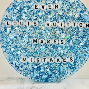 May include: A round, blue glitter coaster with white letters spelling "Even Louis Vuitton Makes Mistakes".