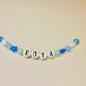 May include: A white beaded garland with blue and white beads and stars, spelling out the name "ROYA".