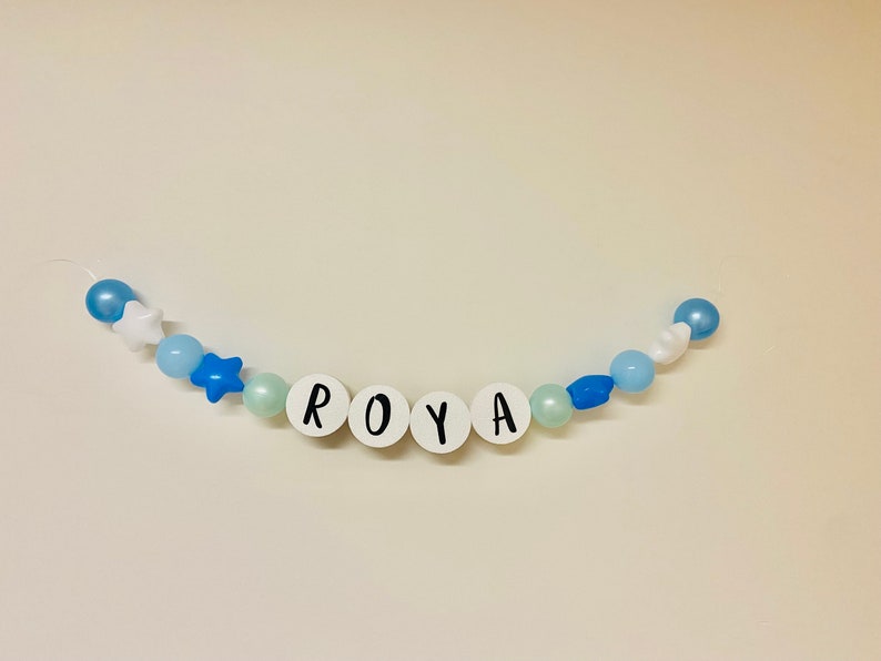 May include: A white beaded garland with blue and white beads and stars, spelling out the name "ROYA".
