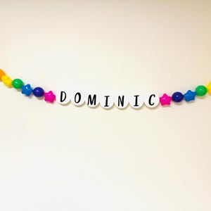 May include: A rainbow coloured beaded garland with white letter beads spelling out the name "DOMINIC".