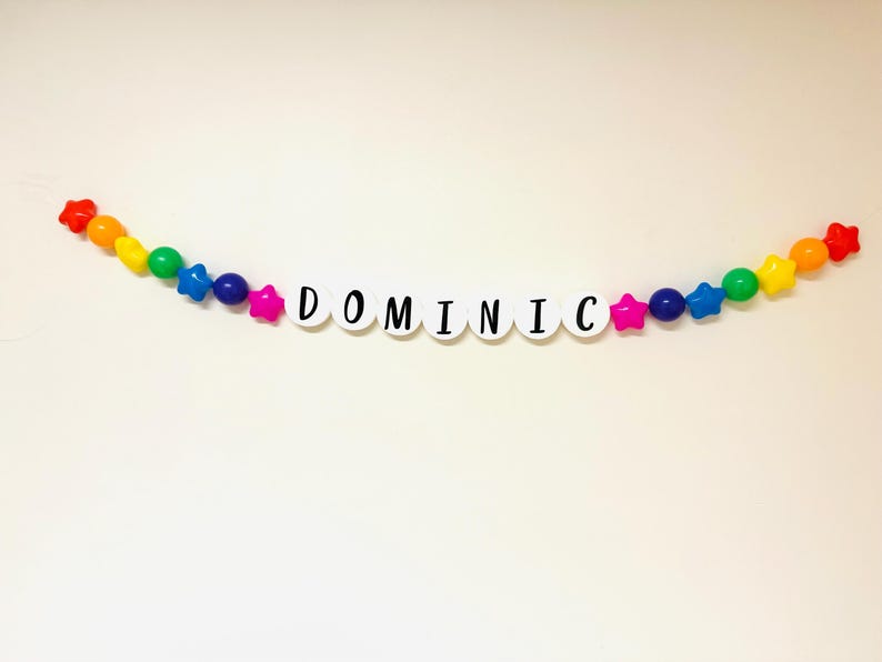 May include: A rainbow coloured beaded garland with white letter beads spelling out the name "DOMINIC".